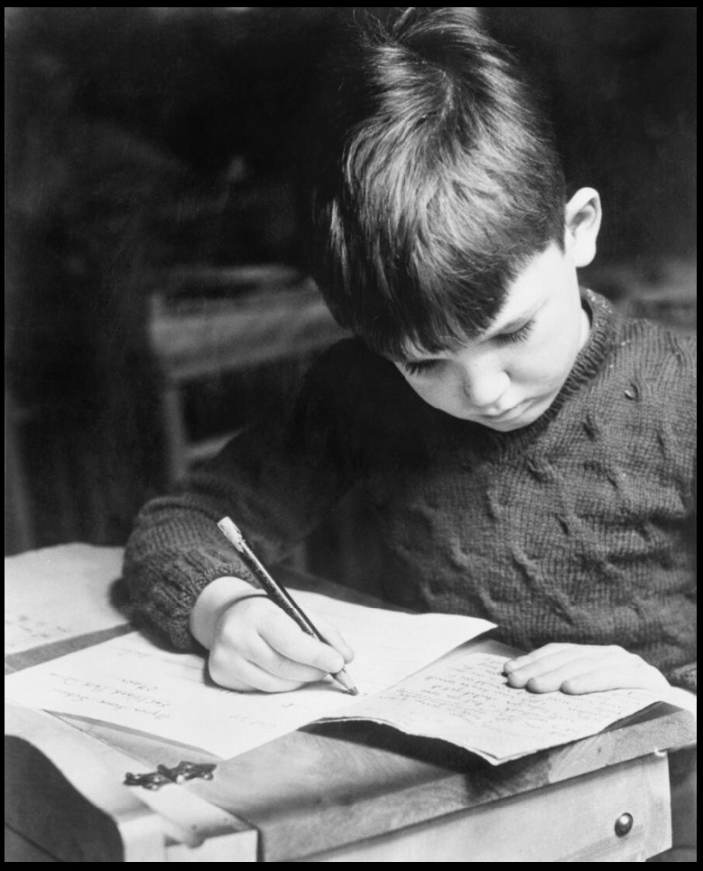 A black and white photo of a child writing at his desk. 