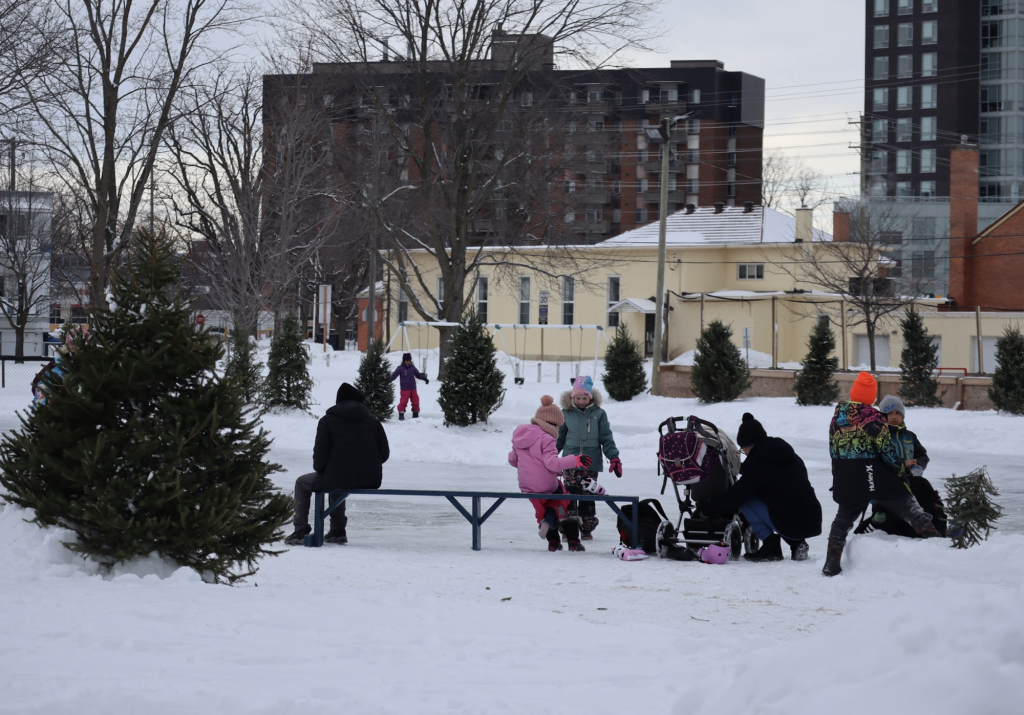 Children lace up their skates at an outdoor rink. Apartments are seen in the distance. 