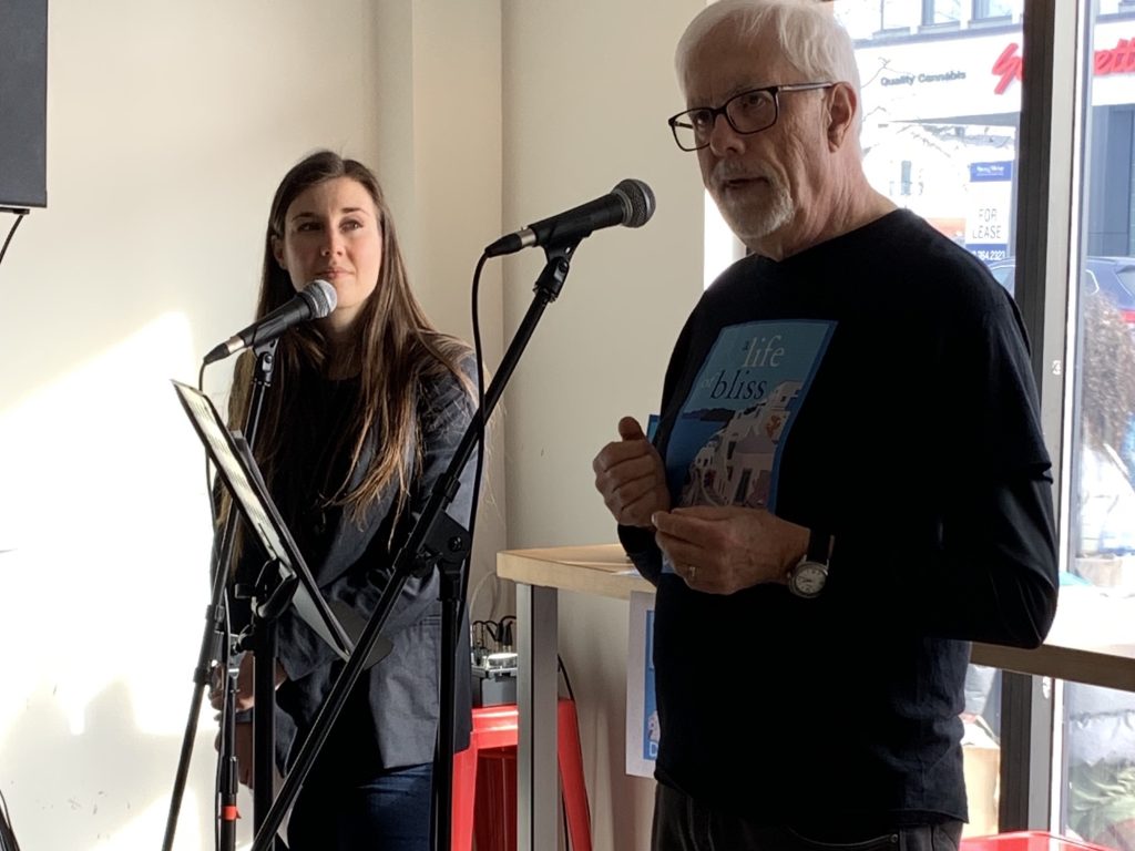 Don speaks into a microphone inside the bagel shop. A woman is seen in the distance. 