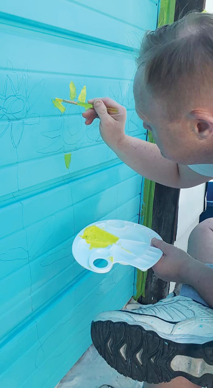 A photo of Paul Knoll painting a garage in Westboro.|A photo of Paul Knoll sitting in front of a garage door mural in Westboro.