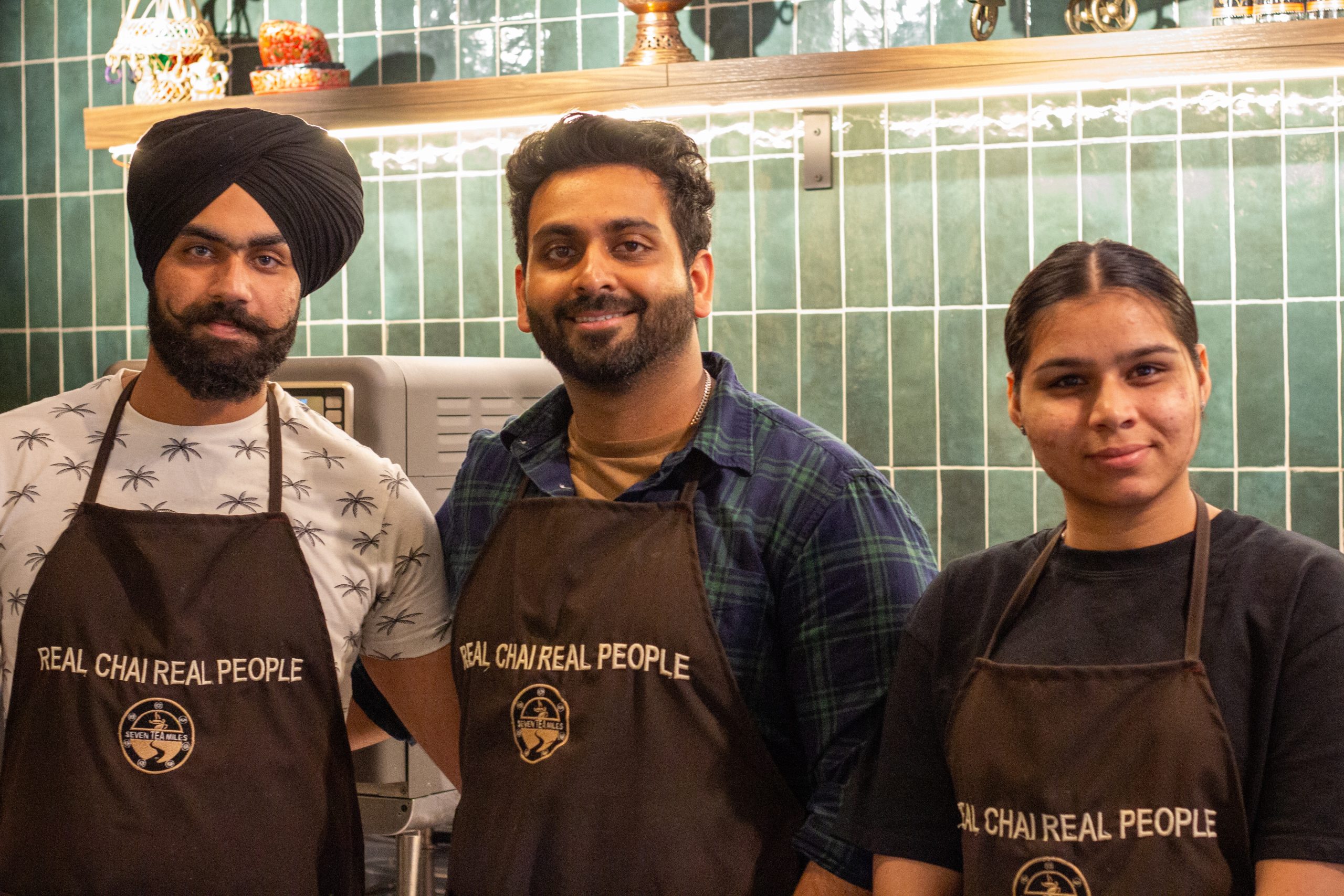 Three people pose for a photograph. Dark green tiling is behind them. 