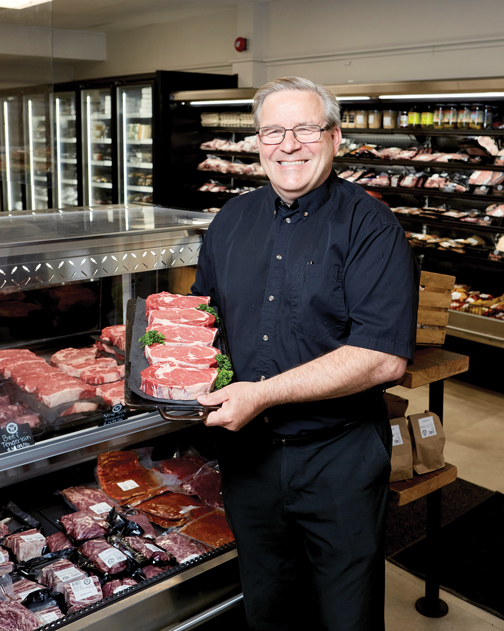 A man dressed in all black holds up beef cuts at a butchery. Behind him