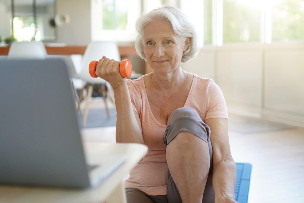 A senior woman sits on a yoga mat and uses an orange hand weight