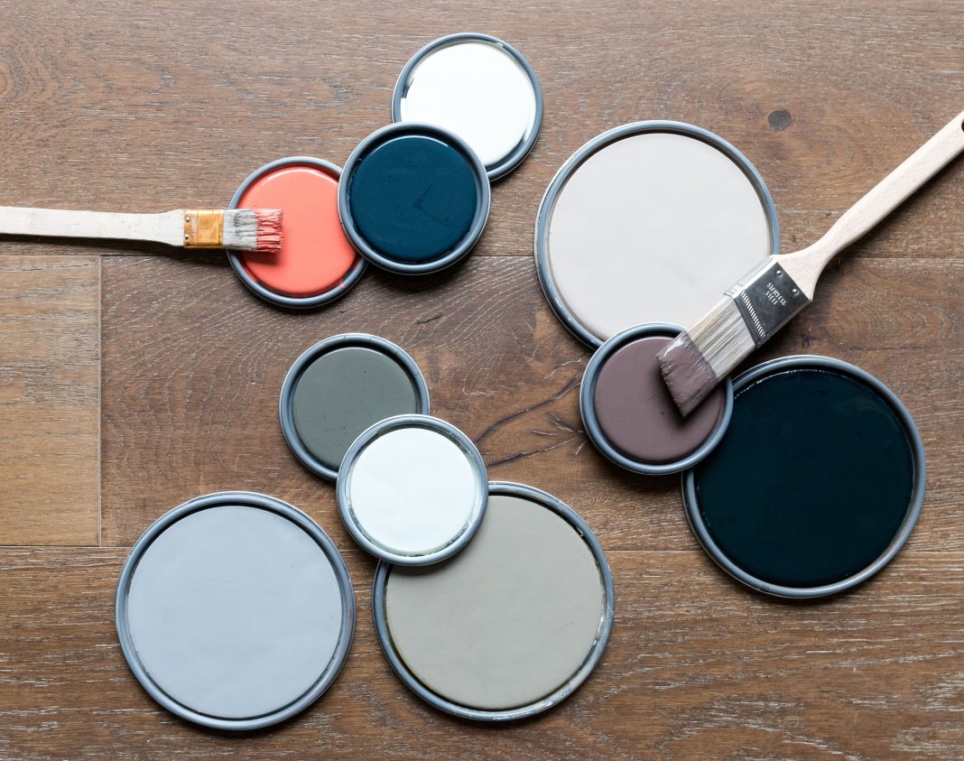 A group of paint can lids and brushes with different paint colours sits on a wooden floor.|