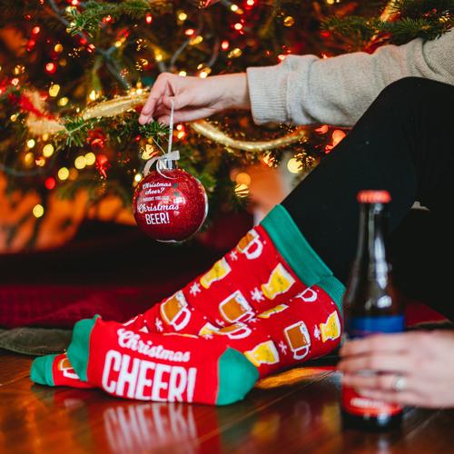 A hand holds a red ornament near a tree while the other hand holds a beer.