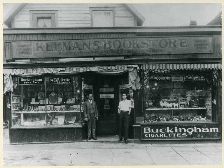 A black and white photo from 1935 showing two men standing outside of Kelmans Bookstore on Wellington West Street|A black and white photo of Fae Taller standing on a snowy Bayview Road in the 1930s in Hintonburg.|A black and white ad from 1922 in the Ottawa Citizen featuring Bodnoff's store|A black and white ad in the Ottawa Citizen from 1930 featuring I.L. Arron Ltd. department store.|A black and white photo of Jacob and Mary Agnes sitting in a delivery truck in 1927.