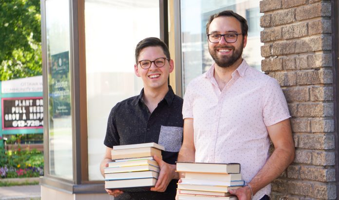 Two men stand side by side holding piles of books outside of The Spaniel's Tale bookstore.|Two men wearing glasses stand next to a blue sign for The Spaniel's Tale bookstore on a sunny day.|A black and white Springer Spaniel dog sits and wears glasses.