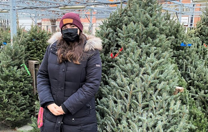 A woman wearing a black coat and black mask stands in front of Christmas trees at Parkdale Market|