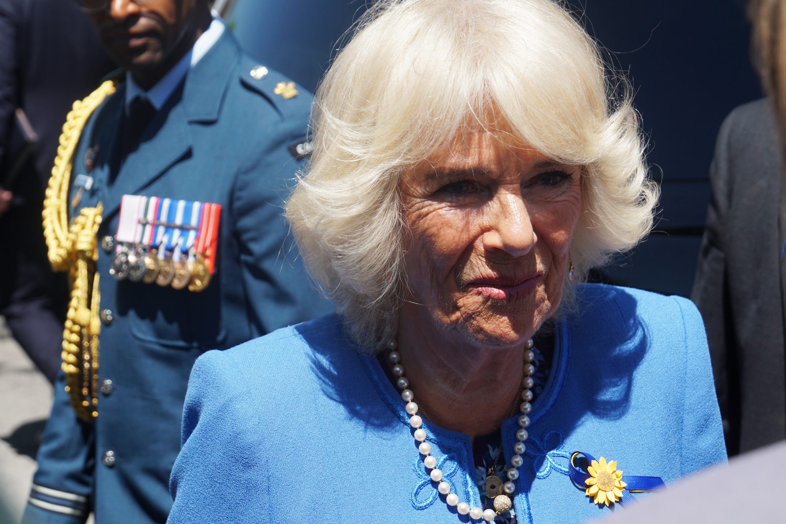 Duchess Camilla is seen wearing a light blue suit with a sunflower pin and a pearl necklace. It is a sunny day and there is a royal guard in uniform behind her.