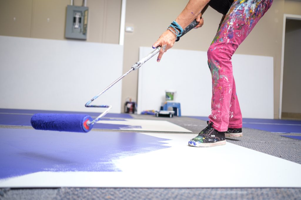 A close up of Claudia Salguero painting a white panel on the floor with blue paint