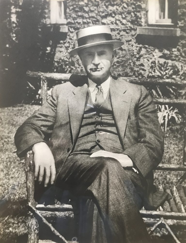 A black and white portrait of a man in a three piece suit and hat sitting on a wooden bench.|||