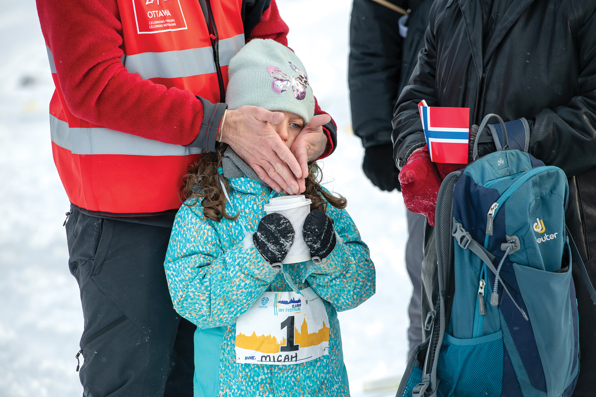 A young girl sips hot chocolate while getting her cheeks warmed up by a pair of hands.