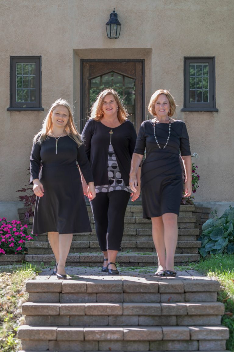 Three women stand in a row wearing black. They are standing on a set of concrete stairs outside.|