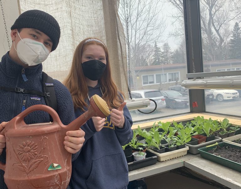 Two students stand in Nepean High School's greenhouse. One carries a watering pot.