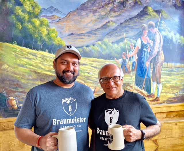 Rohit and Ravi Gupta stand side by side holding beer steins with a colourful mural behind them