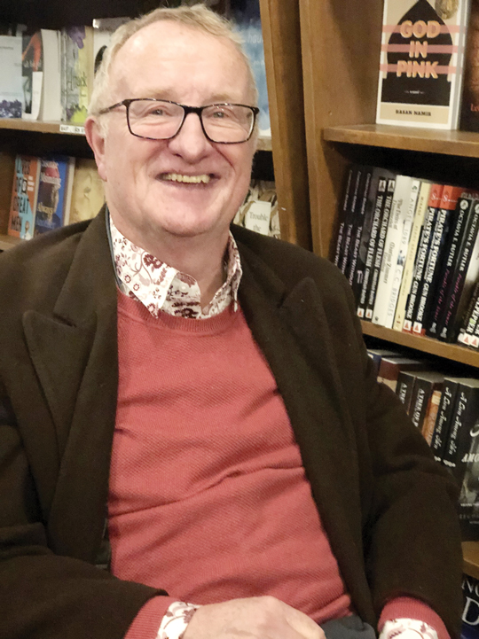 A man wearing a brown jacket and red sweater sits in front of a row of bookshelves.