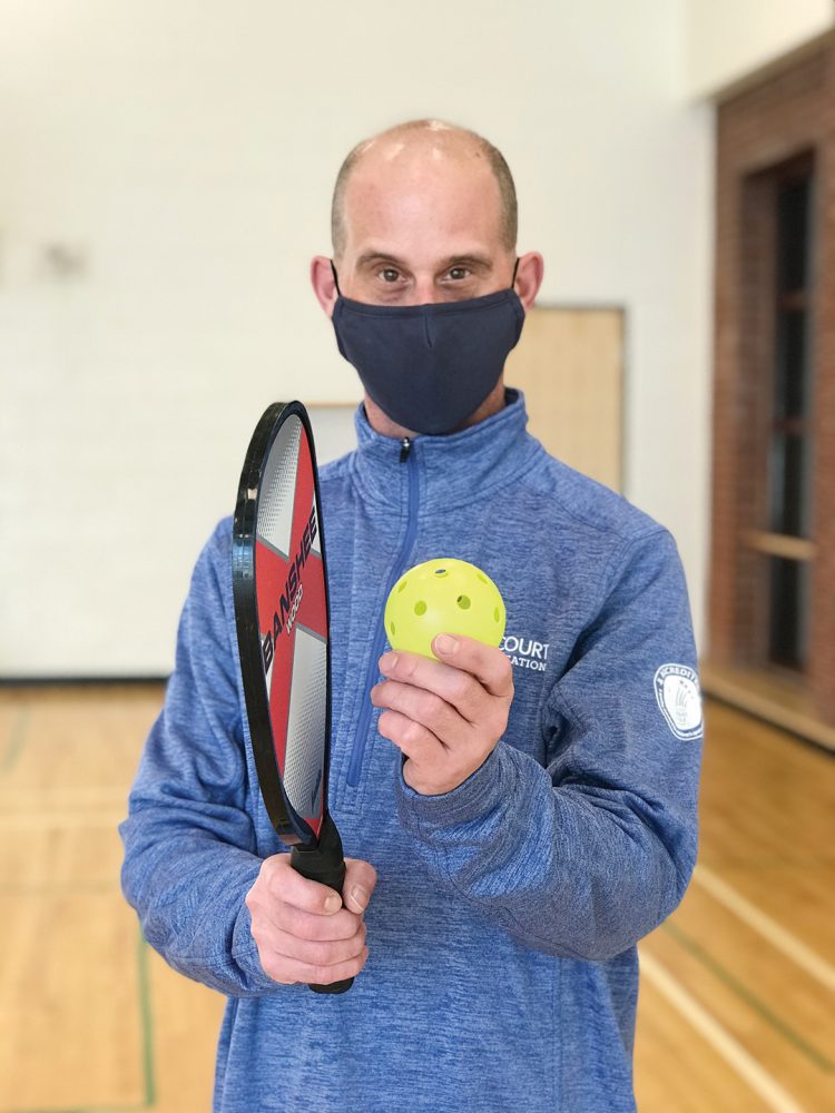 A male instructor in a mask holds a pickleball racket and ball at Dovercourt Recreation Centre