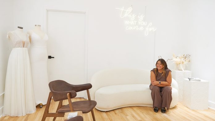 A woman sits on a white couch in a bridal salon with two bridal gowns hanging to the left of her|