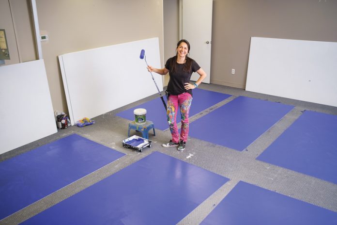 Claudia Salguero stands with a paint roller and blue panels around her on the floor in the middle of a studio space.|A close up of black sneakers and pink pants that are splattered with paint.|A close up of Claudia Salguero painting a blue panel on the floor|A close up of Claudia Salguero painting a white panel on the floor with blue paint