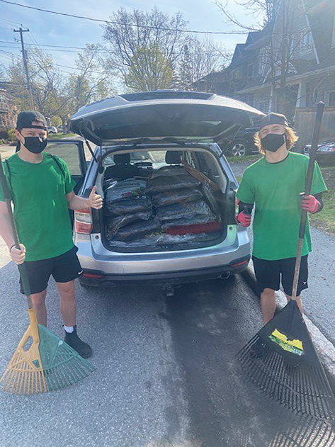 Two teens stand with rakes next to an open trunk of a vehicle with bags of manure and mulch in it|A green and white sign on a lawn that reads 