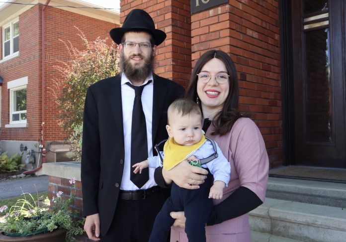 A man wearing a black hat and black suit stands next to a woman wearing a pink dress who holds a newborn boy wearing a yellow bib. There is a brick building behind them.|The exterior of a brick building at 166 Huron Avenue on a sunny day.