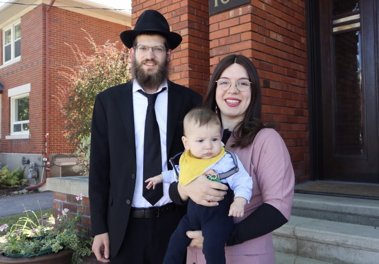 A man wearing a black hat and black suit stands next to a woman wearing a pink dress who holds a newborn boy wearing a yellow bib. There is a brick building behind them.|The exterior of a brick building at 166 Huron Avenue on a sunny day.