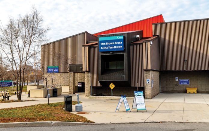 The outside of the main entrance of Tom Brown Arena on a sunny day in Ottawa|Green cots are set up six feet apart on the floor of the Tom Brown Arena Respite Centre|A brightly coloured welcome banner hangs at the Tom Brown Arena Respite Centre