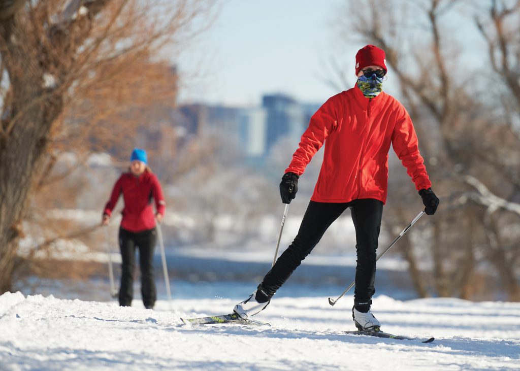 A skier wearing a red jacket, black pants and a face mask is seen up close on the winter trail on a sunny day