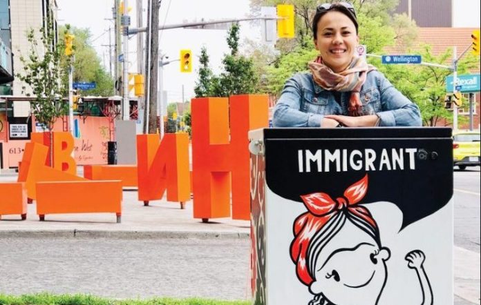 Kseniya Tsoy stands next to a painted Bell Box at the corner of Parkdale and Wellington West. The painting is black and white and shows a woman wearing a red bandana with the word 