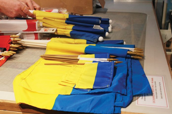 Blue and yellow flags of Ukraine lay flat on a table|A man and woman stand in front of a set of maps on the wall