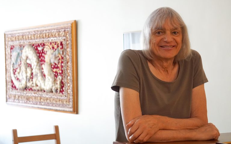 A woman leans against a bookshelf and smiles. Behind her