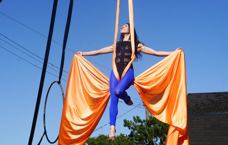 An aerial artist is seen in the air with orange silk ropes. She is wearing blue leggings and a black leotard.|Rows and rows of white and orange pumpkins at the Parkdale Market.|Cardboard baskets filled with apples sit at a table at the Parkdale Market.|A woman is seen in the air performing aerial acts. She is wearing a black leotard and blue skirt and there are orange silk ropes.|A close up of a green wreath with poppies on it. It reads "Her royal majesty Queen Elizabeth II" in a black banner with gold writing.|A group of people does yoga on a lawn on a sunny day in Hintonburg.|The stained glass ceiling at the Tunney's Pasture LRT station on a sunny day.