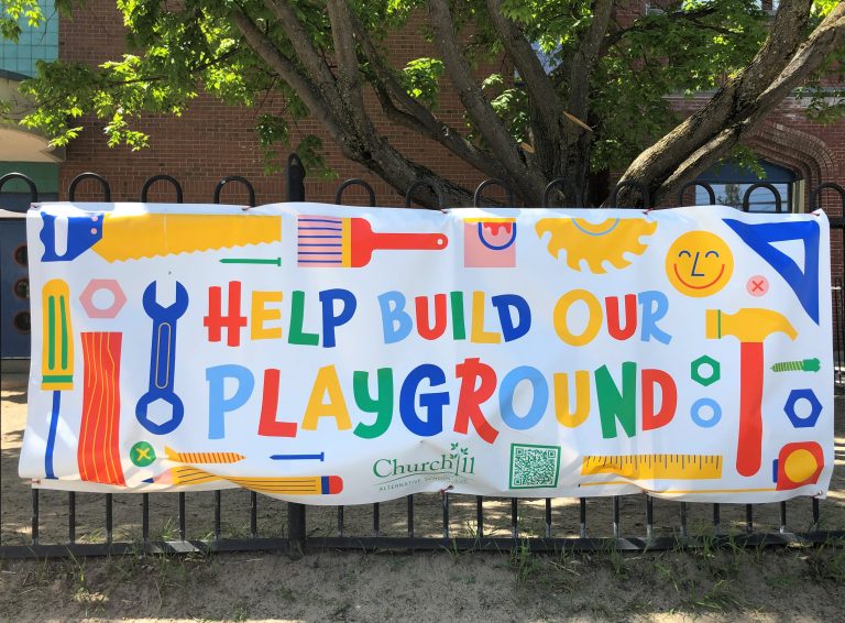 A multicoloured banner outside Churchill Alternative School reads "Help build our playground."|The outside yard for school children at the Churchill Alternative School with the sandy yard and tall tree.