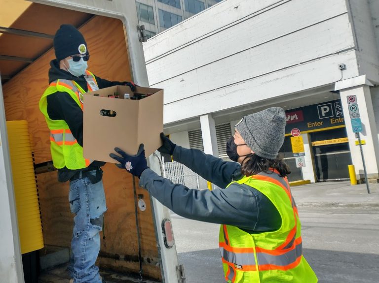 Two men in yellow worker vests and face masks unload the back of a truck. They are passing a cardboard box with bottle in it.|||