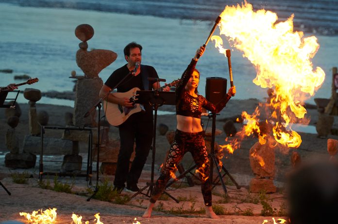 A female fire dancer wearing all black demonstrates a dance with fire-lit batons on a beach at dusk. There is a man playing guitar behind her.||||||||||||