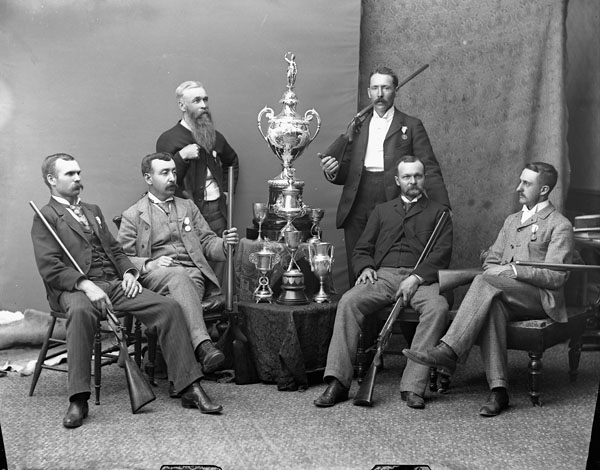 A black and white photo of a group of six men holding guns. There is a stack of trophies in the middle of them. The photo is from the late 1800s.|||||