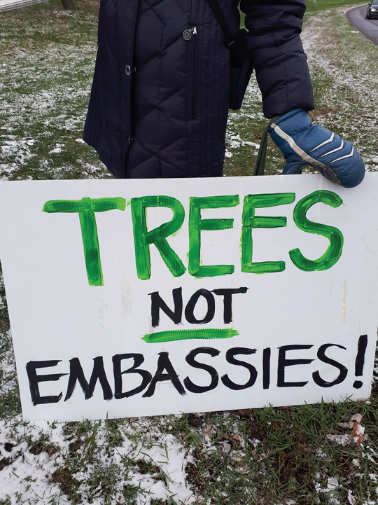 A white protest sign is held up by a gloved hand. The sign reads "trees not embassies" in green and black lettering.|An open field in Mechanicsville is seen with a row of trees in the distance