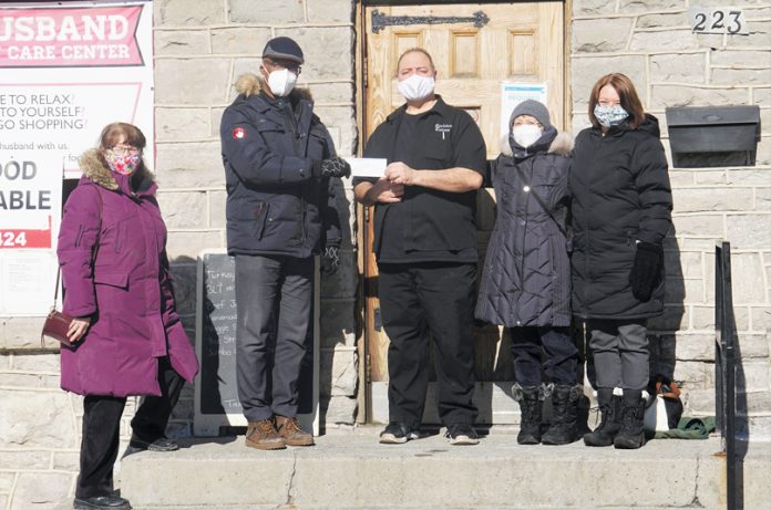 A group of five people stand on the steps of Carlton Tavern on a sunny day in winter. Two of them are holding a white cheque.|