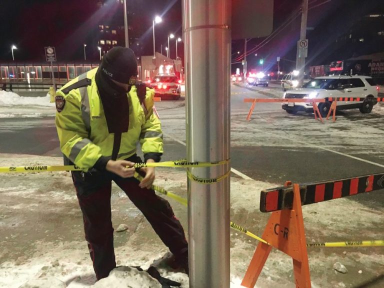 A police officer stands near a pole at the Westboro bus station and tapes off an accident scene|The Westboro bus station is seen from a distance with two reporters on site three days after the crash