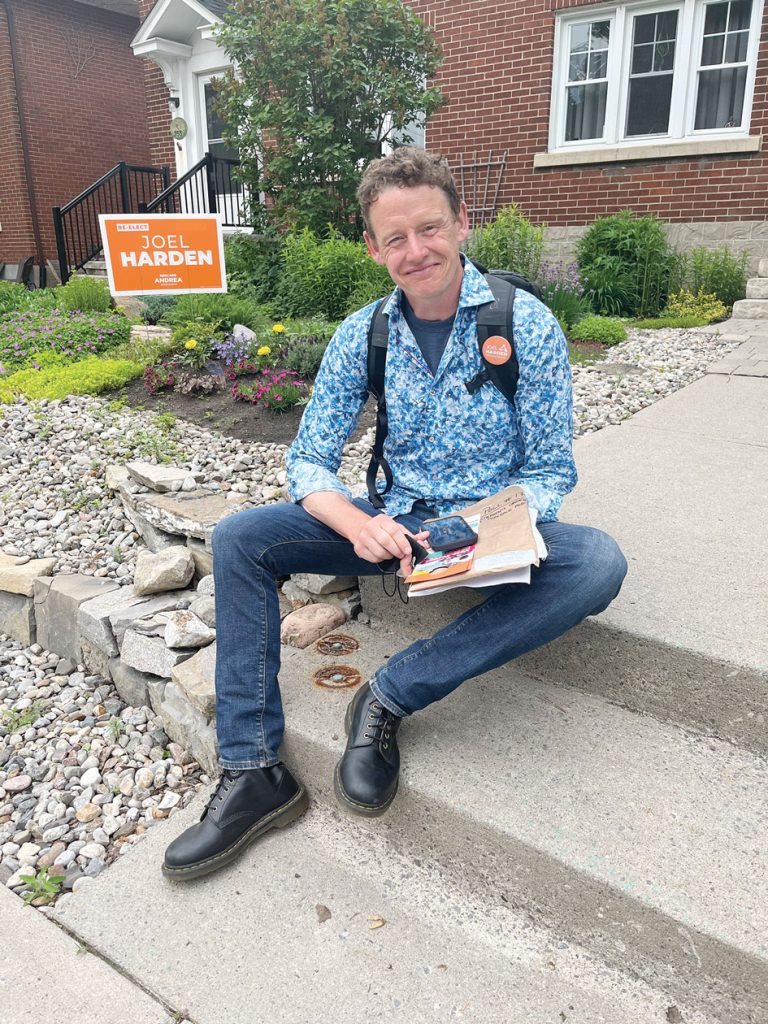 Joel Harden sits on a set of steps wearing blue jeans and a long blue shirt. He is holding flyers and there is an orange Joel Harden NDP sign behind him.|Joel Harden wears a mask and talks to voters at a campaign event outdoors in spring 2022.