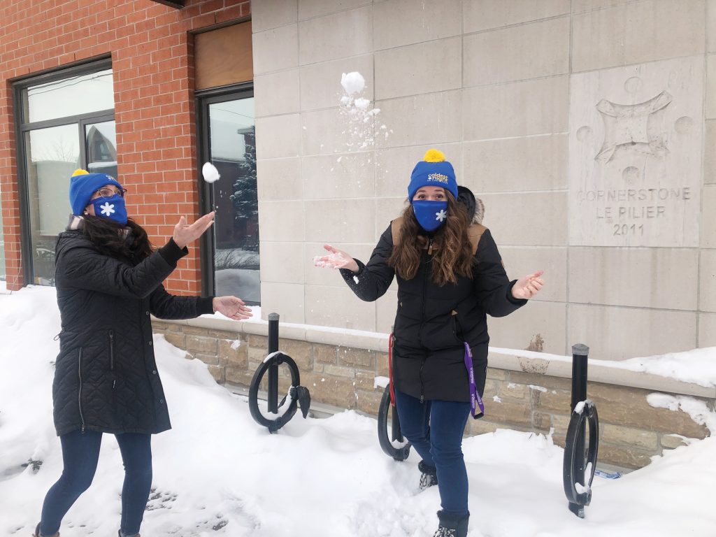 Two Cornerstone staff stand in front of the beige and brick building wearing blue masks and toques and toss snow in the air