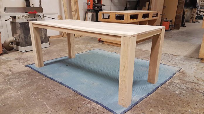 A wooden dining table is seen in the middle of a woodshop.|A professional photo of a man wearing a red t-shirt that says 