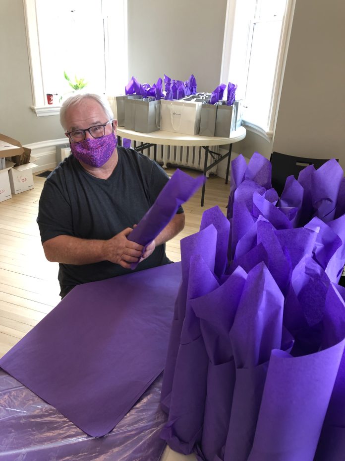 A man wearing a purple face mask fills a table of gift bags with bright purple tissue paper.||