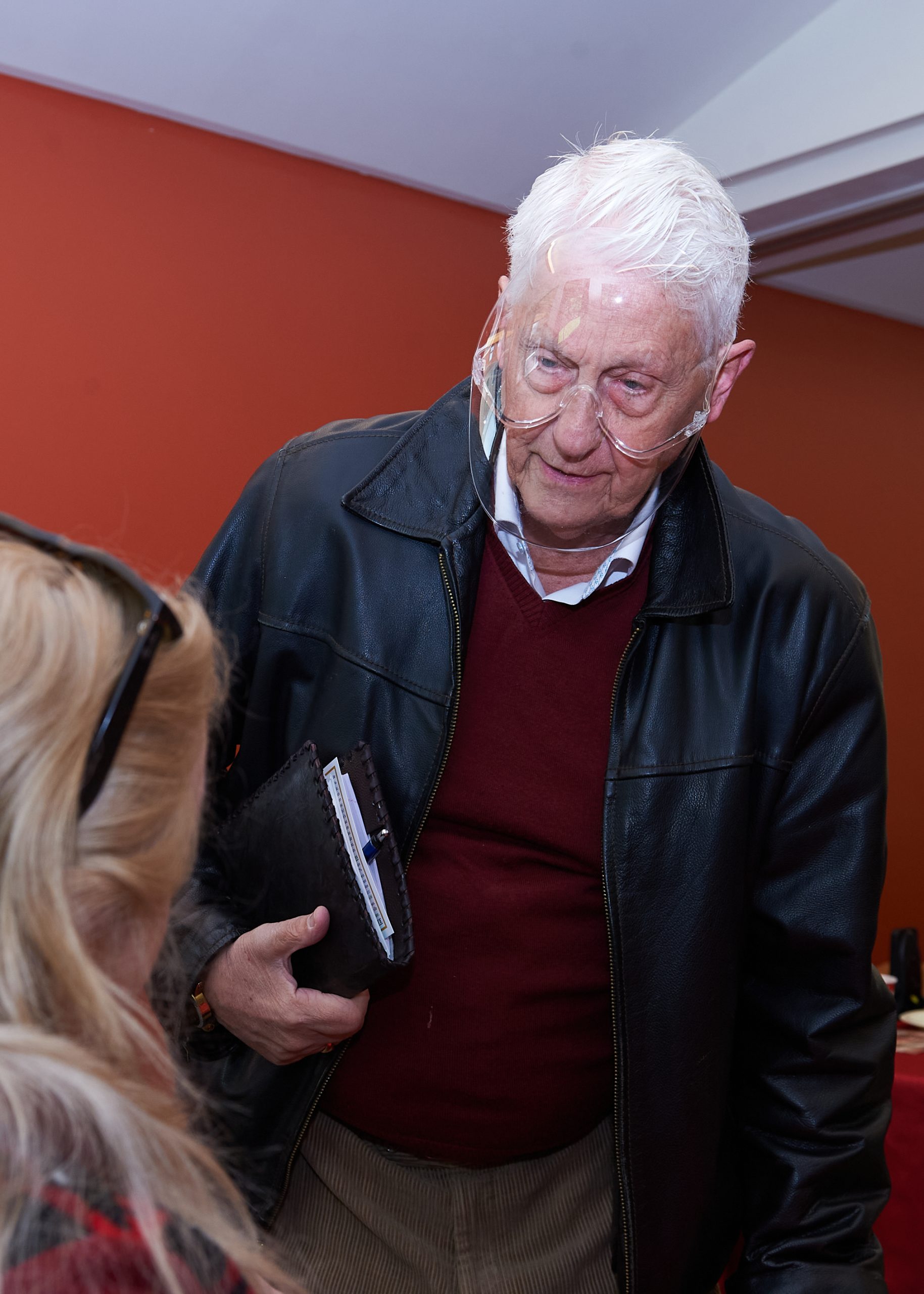 An older man wearing a black jacket, red sweater and face shield talks to a woman with blonde hair.