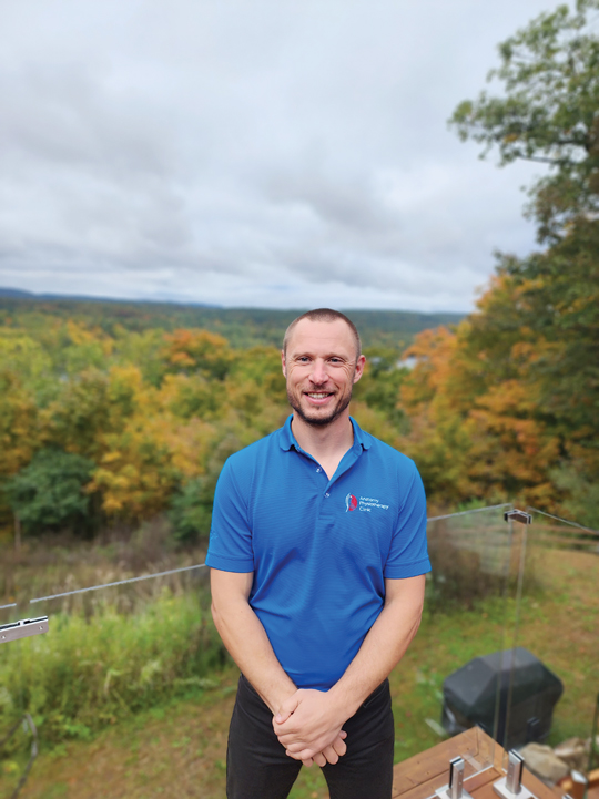 A man wearing a short sleeve blue shirt stands with his hands folded in front of him. There is a fall Ottawa forest behind him.