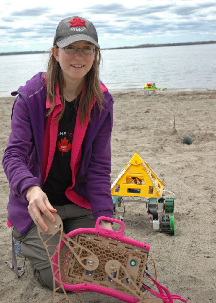 Erin demonstrates her prototype scissor lift beach cleaning bot with her two other bots behind her. Erin designed and constructed all three bots with the help of a 3D printer.