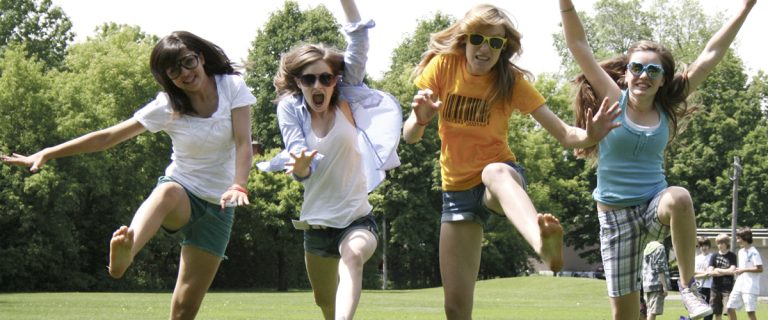 |Four teenage girls jump in the air on a sunny day on a grass field.