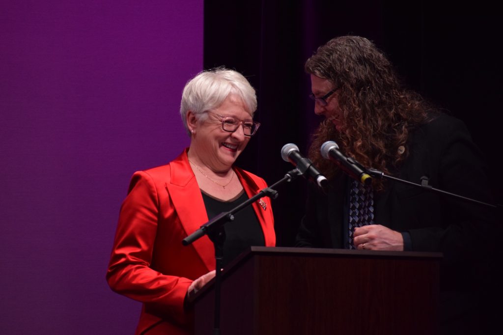 Edith wearing a bright red jacket stands at a podium. 