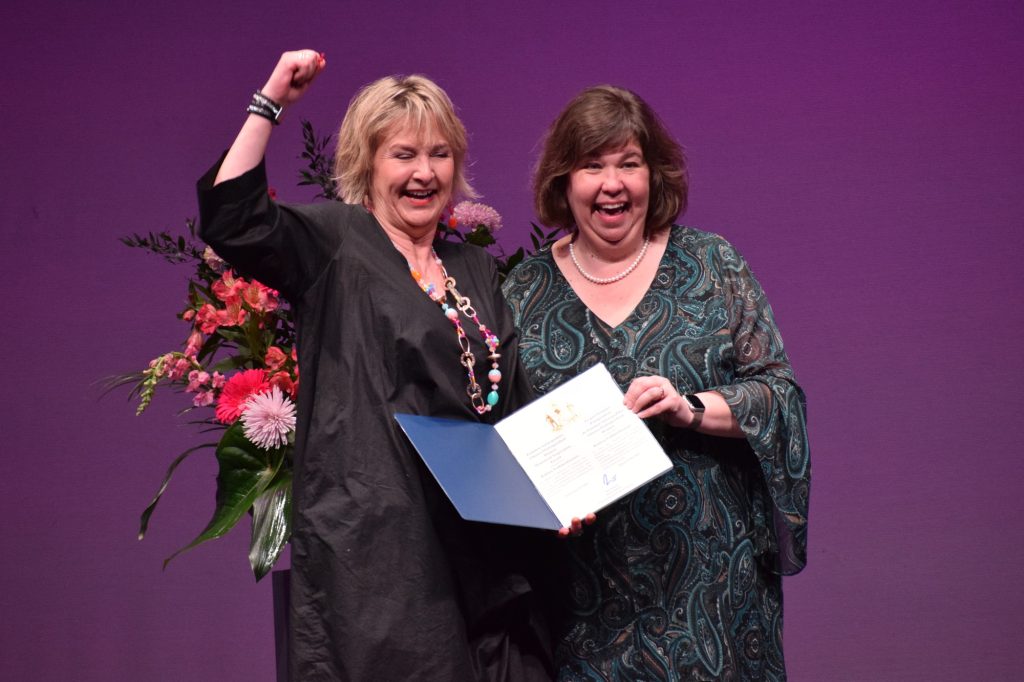 Bettina holds her fist to the air as she receives the certificate from Anita. 