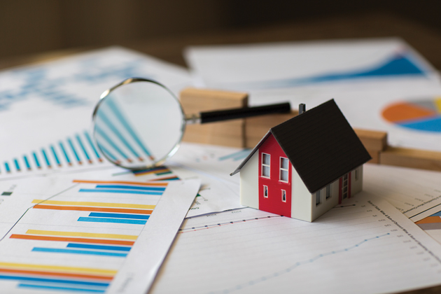 A small white and red toy house sits atop a pile of bar charts and papers. Next to it